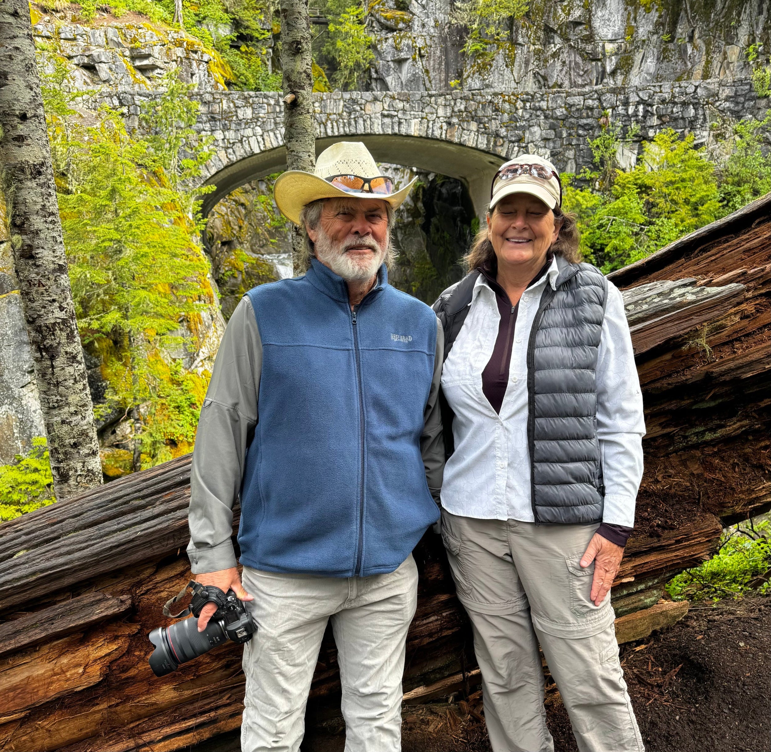 Chuck and Carrie standing in a forested area with a stone arch bridge in the background.